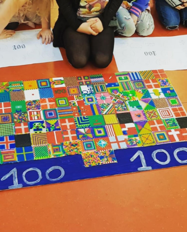 Billedet viser et barn, der knæler ved siden af et værk lavet af klassen, som er fyldt med 100 små firkanter med flag, lavet af perler