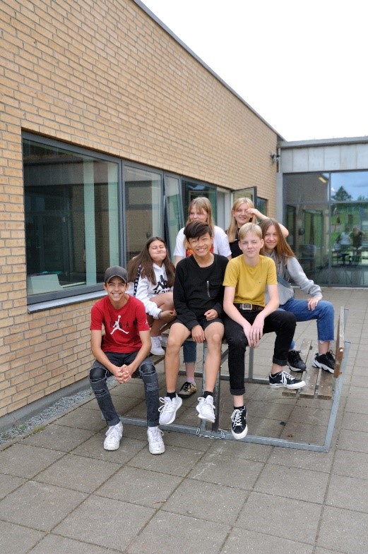 Billedet viser 7 glade og smilende skoleelever, der sidder på et bord uden for en klasselokale på afdeling Fussingsvej