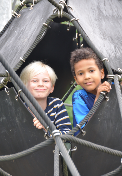 Billedet viser to drenge, der kigger ud af et hul i et klatrestativ og smiler